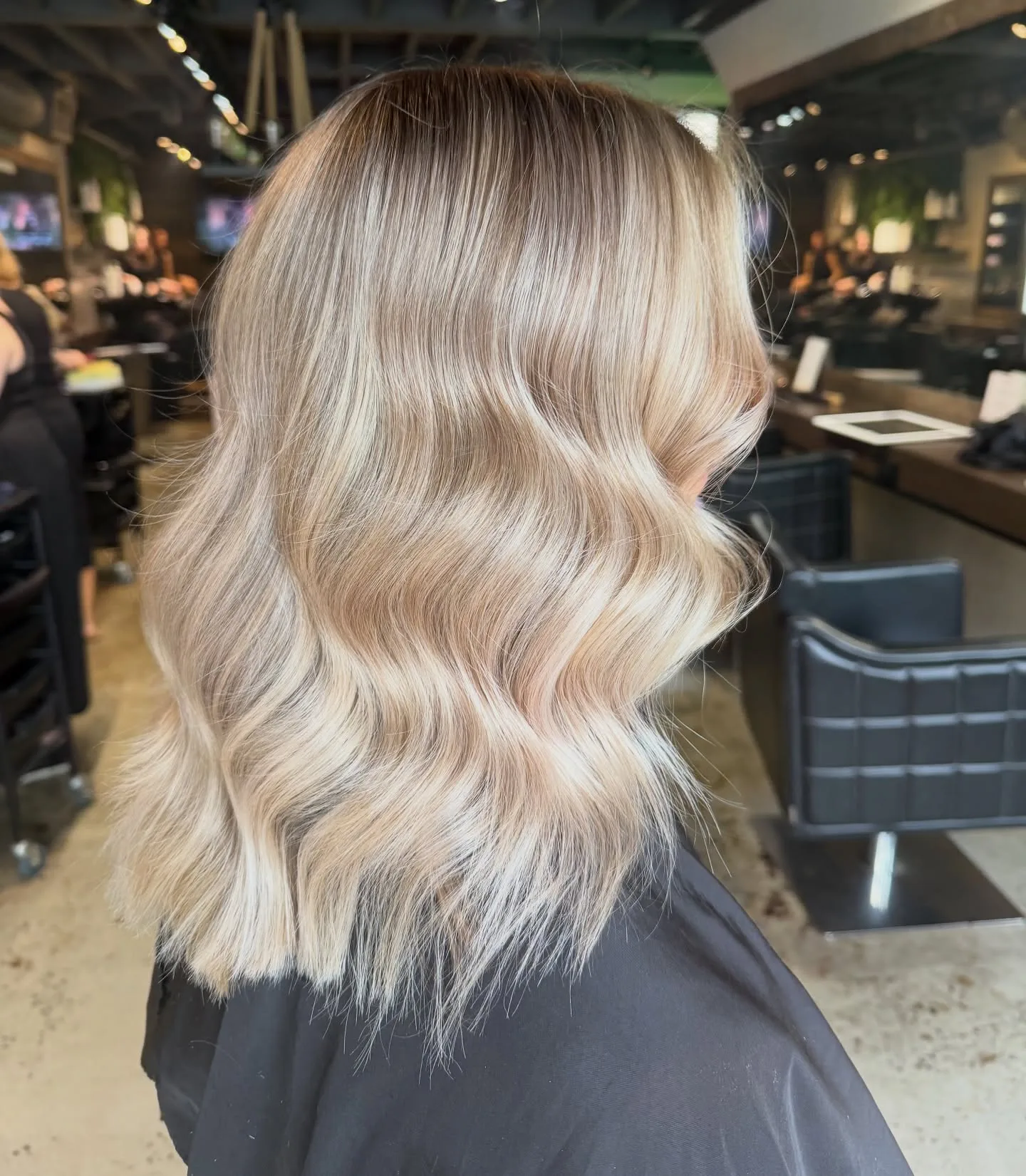 Lived-in blonde balayage mid-length waves, rear-side view — black chairs and mirror in salon