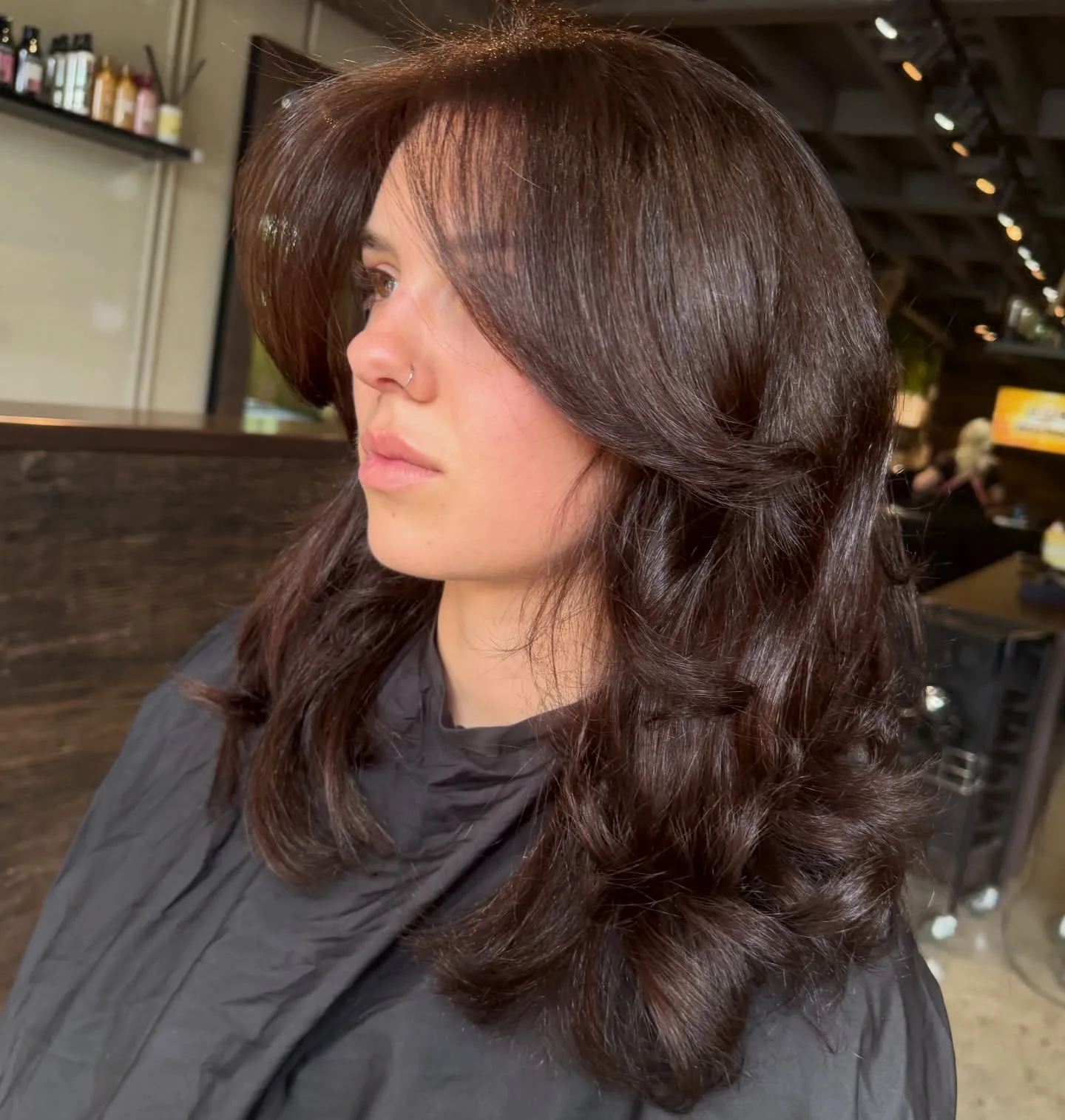 Brunette curtain-bang bouncy blow-dry, client profile — product shelving behind