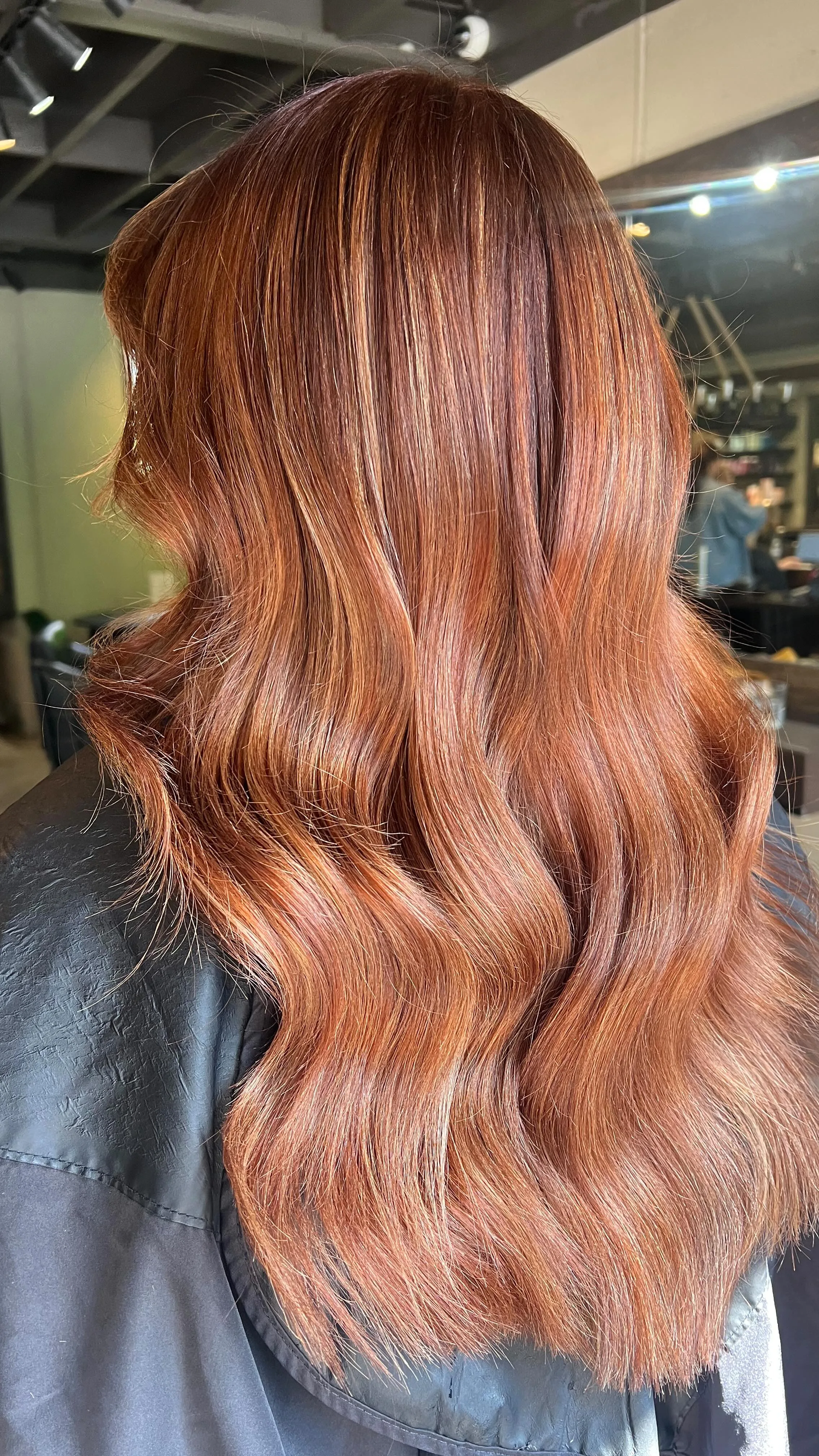 Vivid copper-red long wavy balayage, rear view, warm salon interior