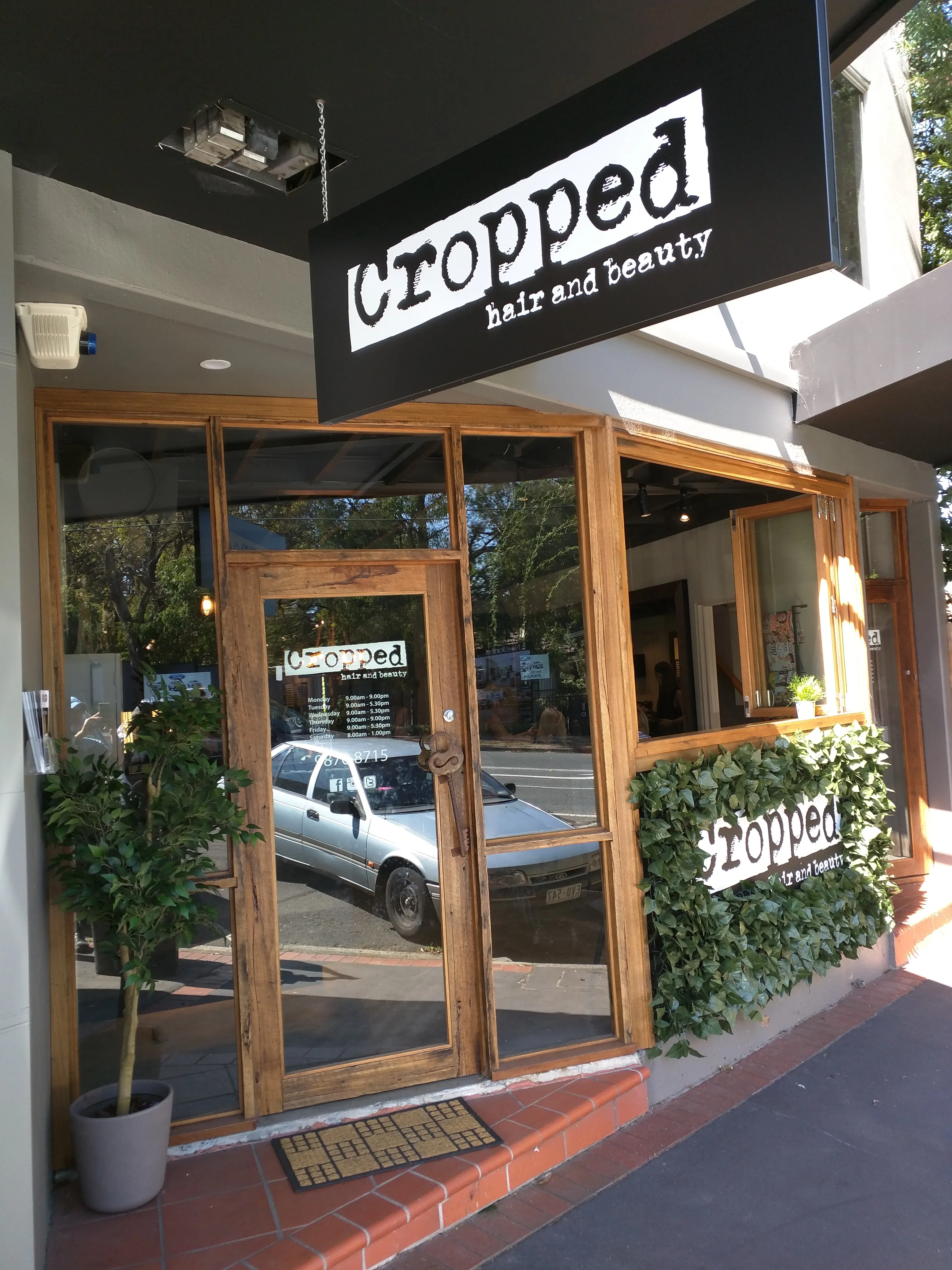 Cropped Hair and Beauty daytime shopfront — black awning wordmark plaque, ivy-hedge sign, Loughnan Road Ringwood